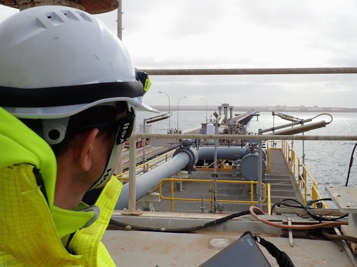 A safety engineer in a hard hat and neon jacket observes industrial equipment and pipelines leading to the water's edge.
